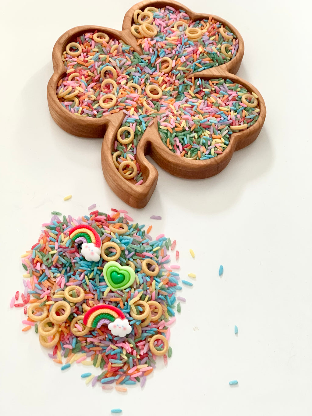 Wooden shamrock tray filled with multicolored rice, small ring pieces, and a pile of colorful rice with rainbow and heart buttons nearby