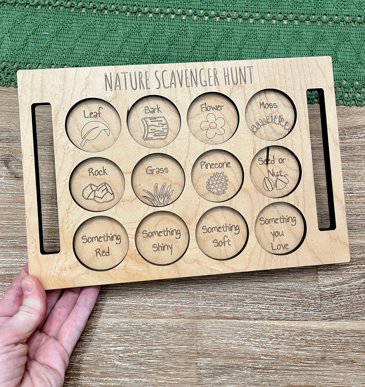 Natural wooden Nature Scavenger Hunt board with engraved circles and nature-themed words and illustrations held by hand on wood surface