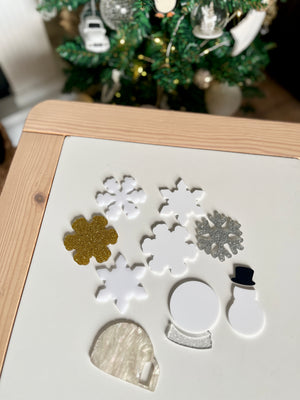 Various white, gold, and silver acrylic holiday shapes including snowflakes, a snow globe, a snowman, and an igloo on wooden table top