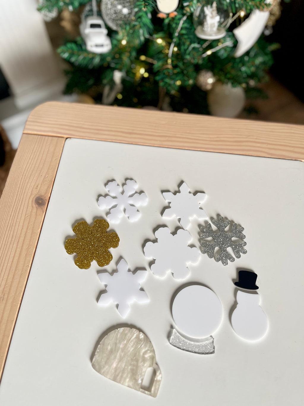 Various white, gold, and silver acrylic holiday shapes including snowflakes, a snow globe, a snowman, and an igloo on wooden table top