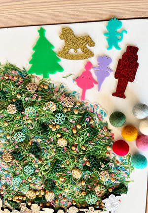Colorful holiday-themed acrylic cutouts including tree, rocking horse, nutcracker, and figurines above mixed sensory materials with felt balls and wooden shapes