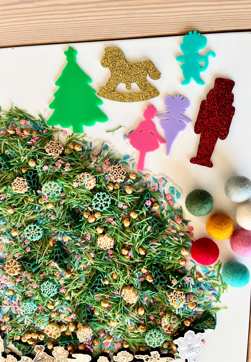 Colorful holiday-themed acrylic cutouts including tree, rocking horse, nutcracker, and figurines above mixed sensory materials with felt balls and wooden shapes