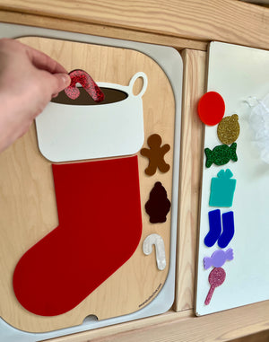 Hand placing a red candy cane shape into a festive stocking cutout on a wooden activity board with holiday shapes nearby