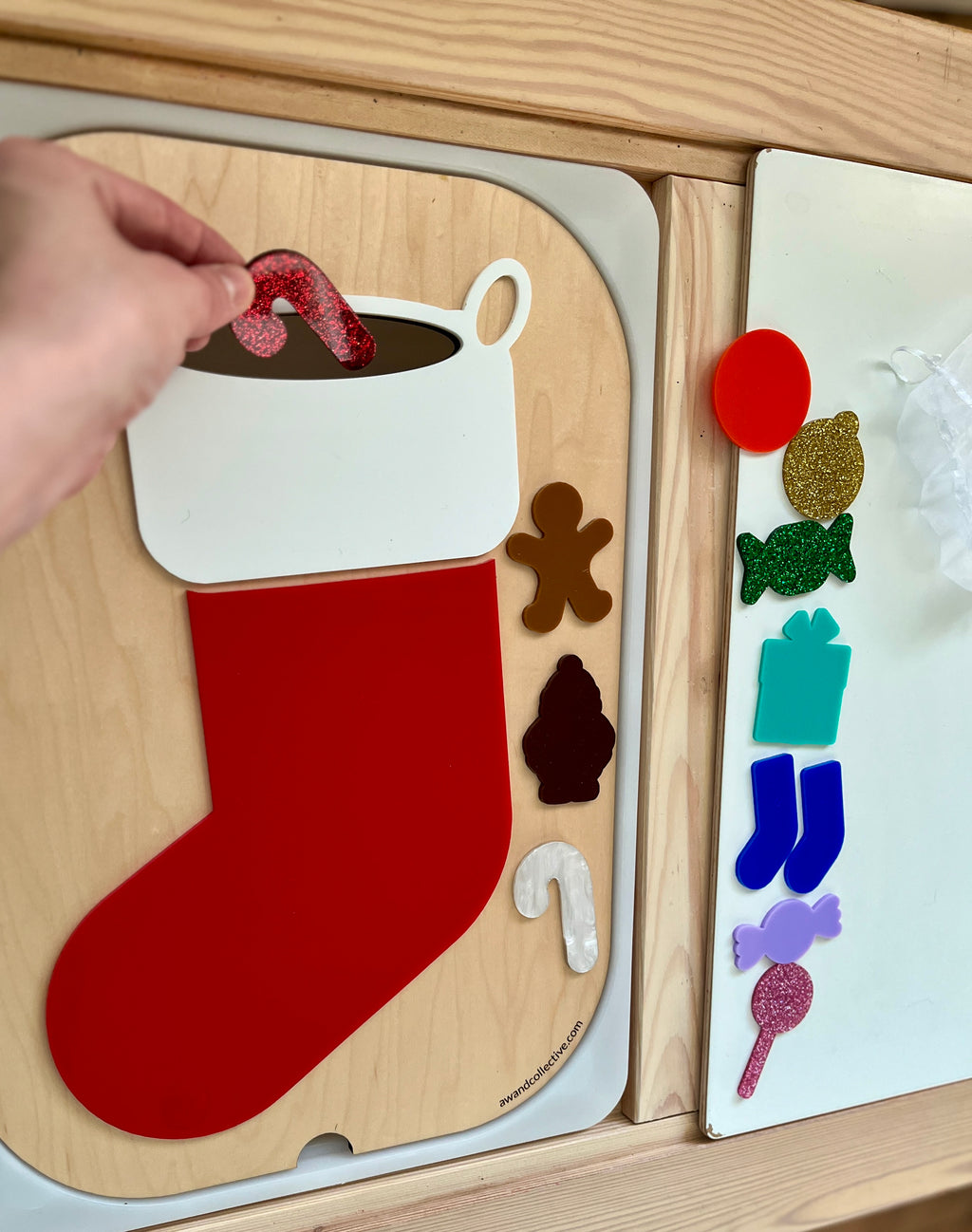 Hand placing a red candy cane shape into a festive stocking cutout on a wooden activity board with holiday shapes nearby