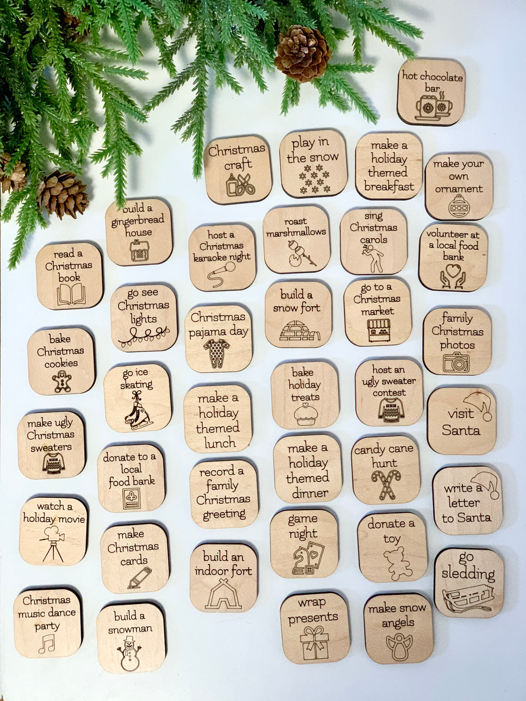 Wooden Christmas activity tokens with holiday-themed illustrations arranged on white surface with pine branches and pinecones nearby