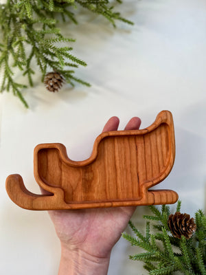 Hand holding a smooth wooden tray carved in the shape of a sleigh with pine branches and cones nearby