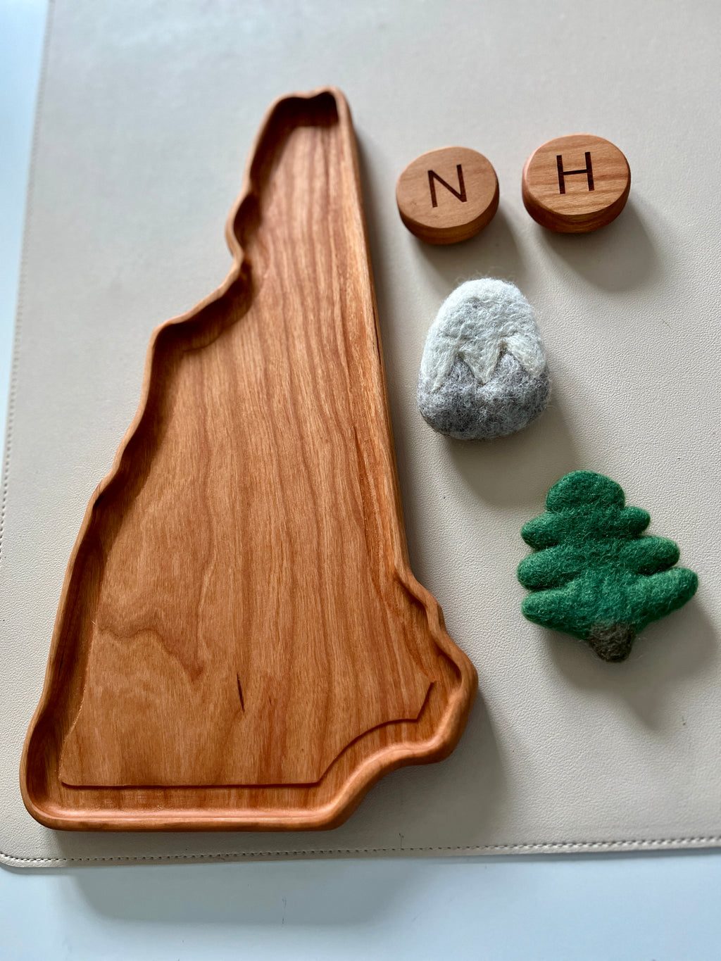 Wooden tray shaped like a state silhouette with felt mountain and tree toys and two wooden letter blocks on a white surface