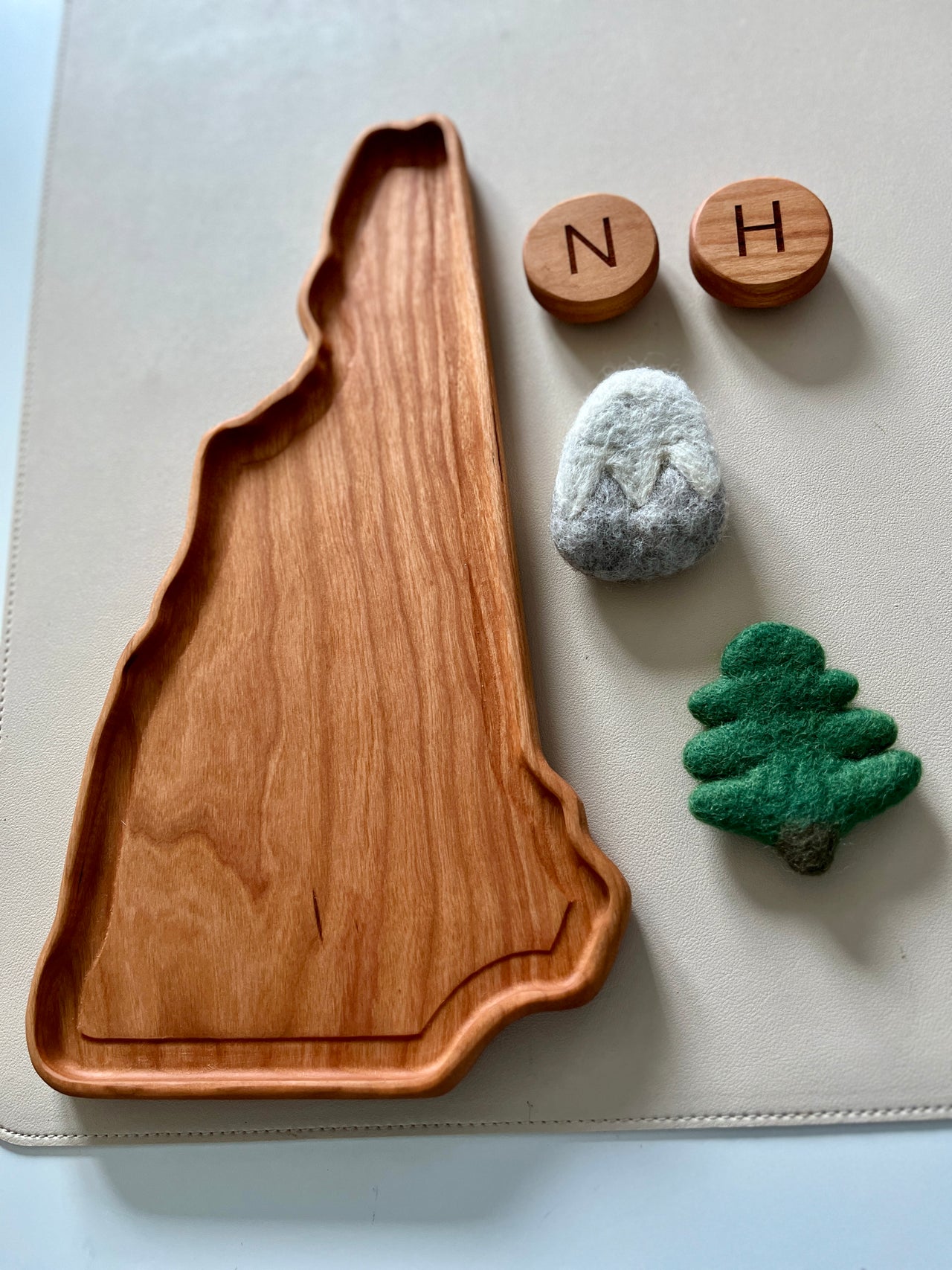 Wooden tray shaped like a state silhouette with felt mountain and tree toys and two wooden letter blocks on a white surface