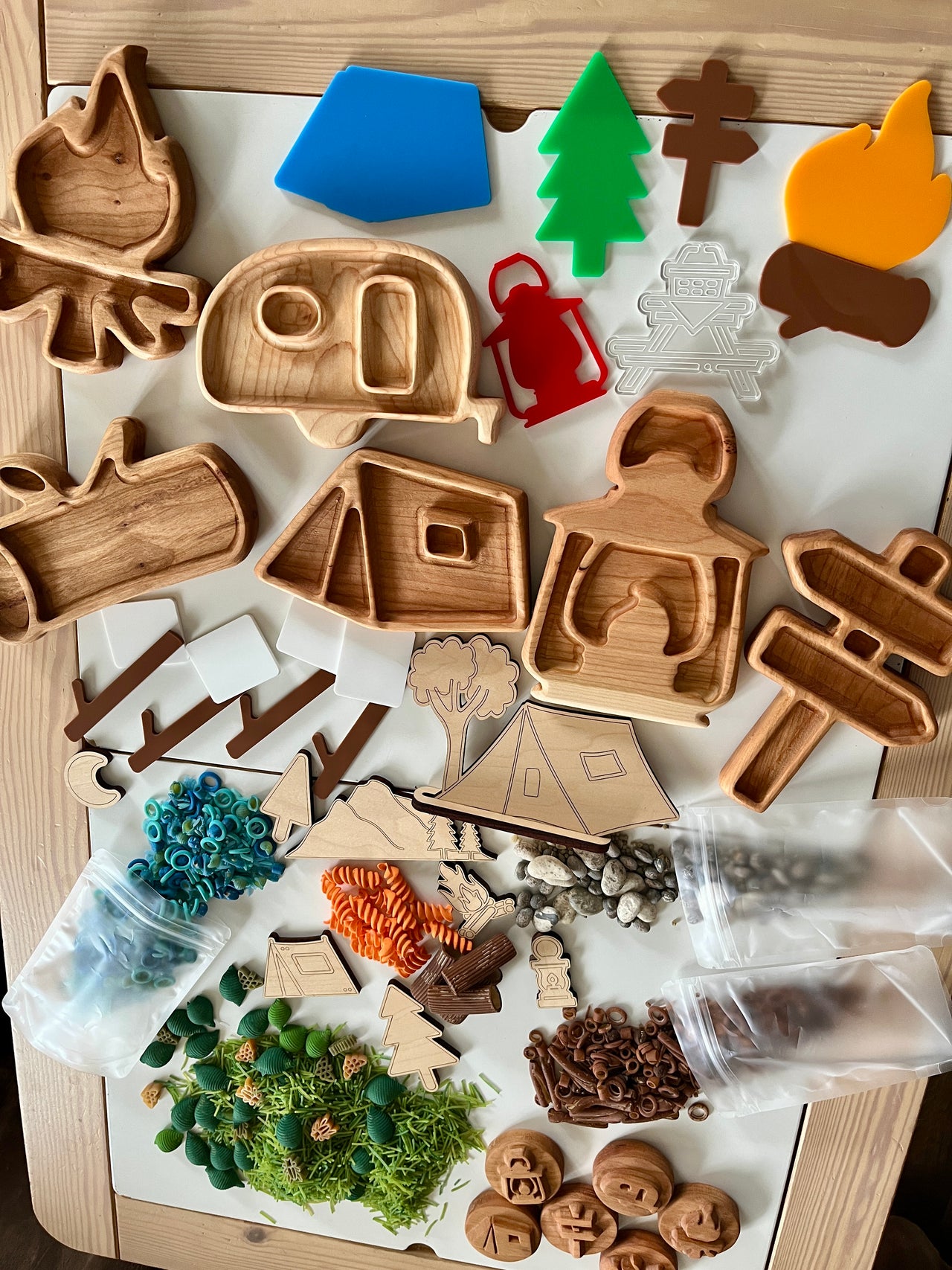 Wooden camping-themed trays and cutouts including tent, camper, lantern, campfire, directional signs, green tree, and other nature elements on table
