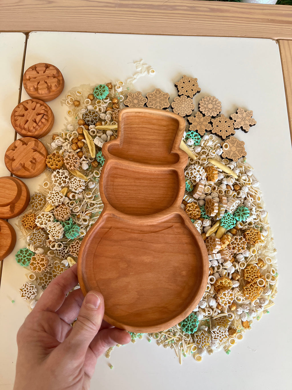 Hand holding wooden snowman-shaped tray above mixed natural and colored sensory materials on white surface