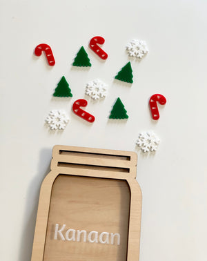 Light wooden jar-shaped frame with the name Kanaan and scattered red candy canes, green Christmas trees, and white snowflake tokens above on a white surface
