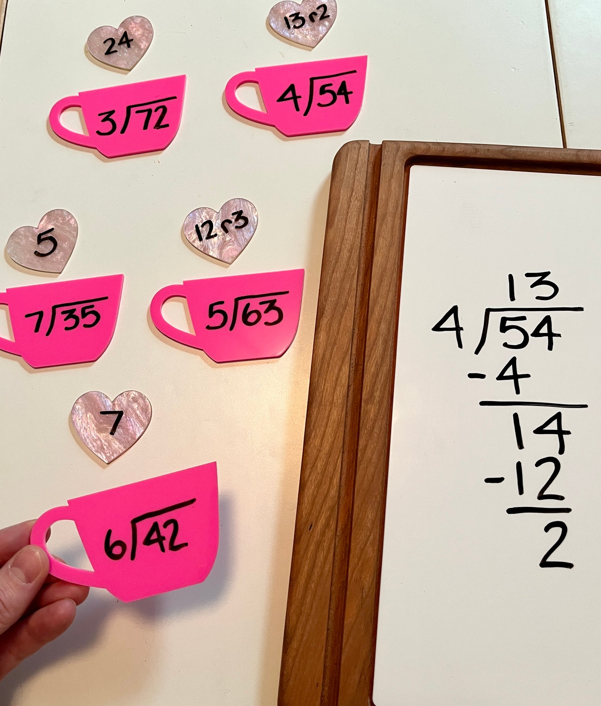 Mathematical equations on pink dry erase shaped like teacups and hearts, with a whiteboard in a wooden tray in the background with the equations on it. 