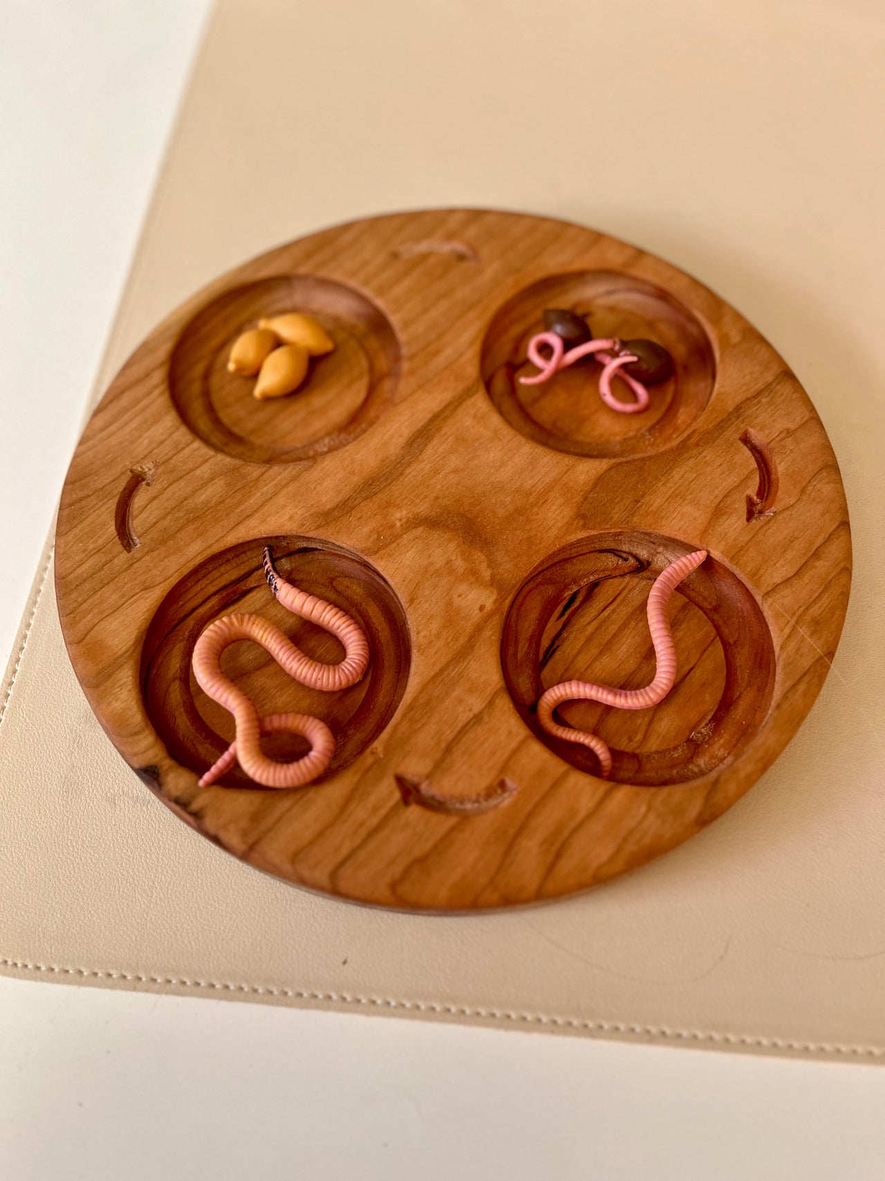 Round wooden sorting tray with four compartments holding pink plastic worms and yellow plastic seeds on a light surface