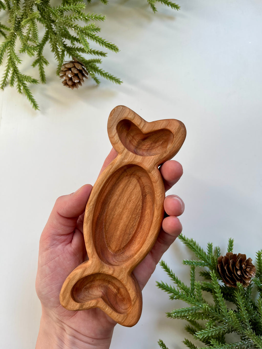 Hand holding a carved wooden tray shaped like a fish with pine branches and pine cones on white background