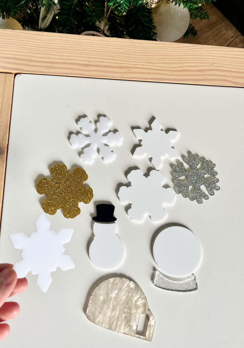 Hand holding white snowflake shape among white, gold, and silver holiday-themed acrylic shapes on table