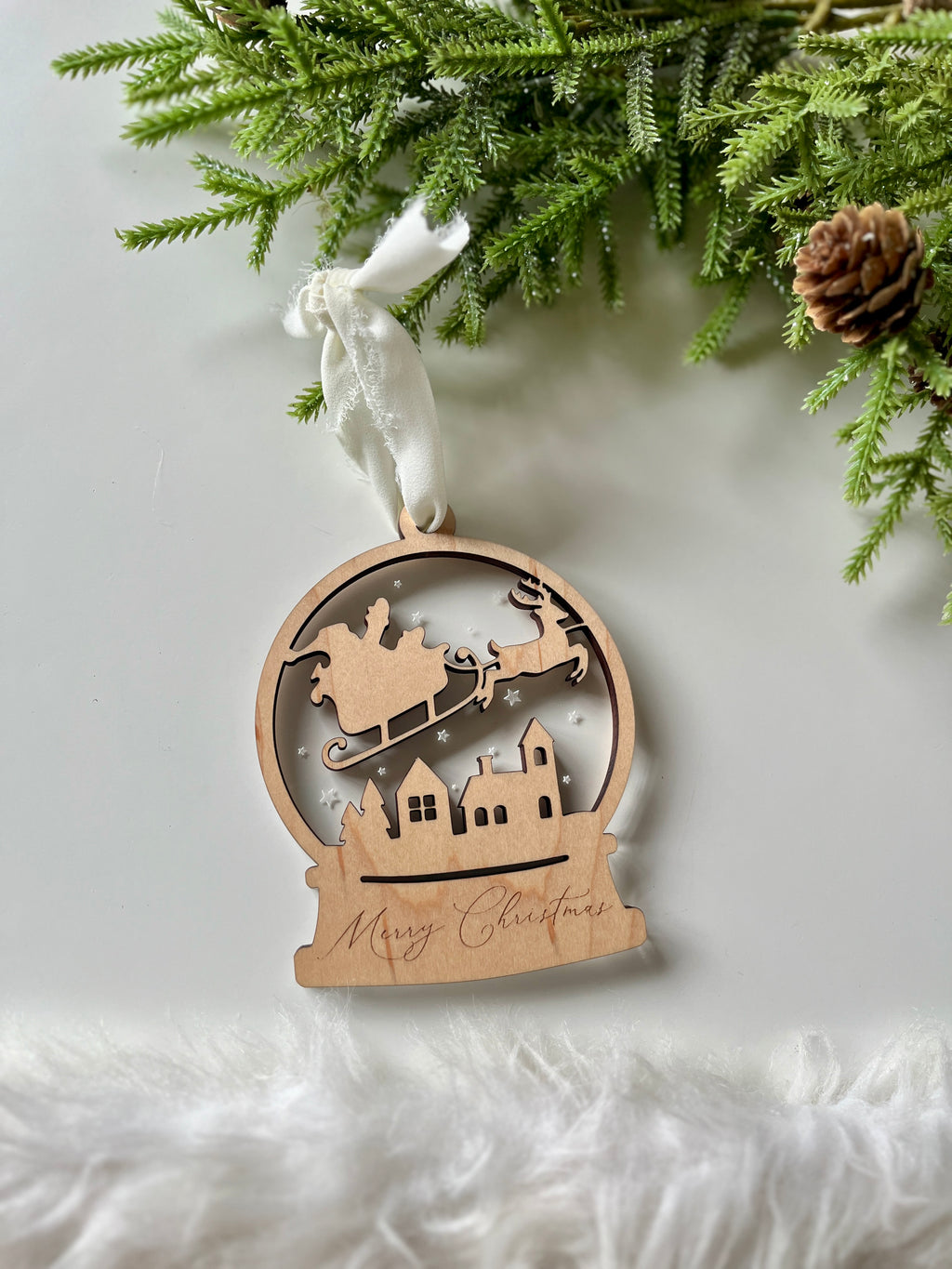 Wooden Christmas ornament featuring Santa in sleigh over village silhouette with "Merry Christmas" engraving and white ribbon