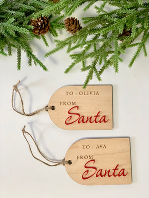 Two rectangular wooden Christmas gift tags with "To" and "From Santa" engraved and tied with twine on white surface