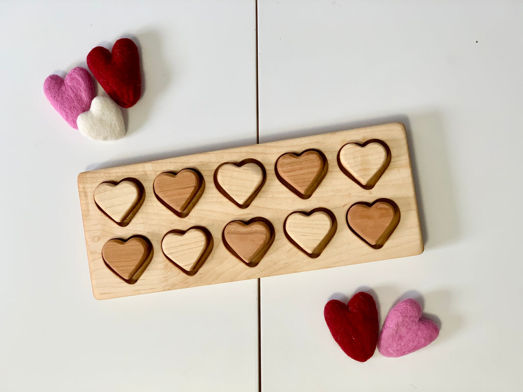 Wooden heart puzzle with alternating light and dark hearts and felt hearts in red, pink, and white nearby on white surface