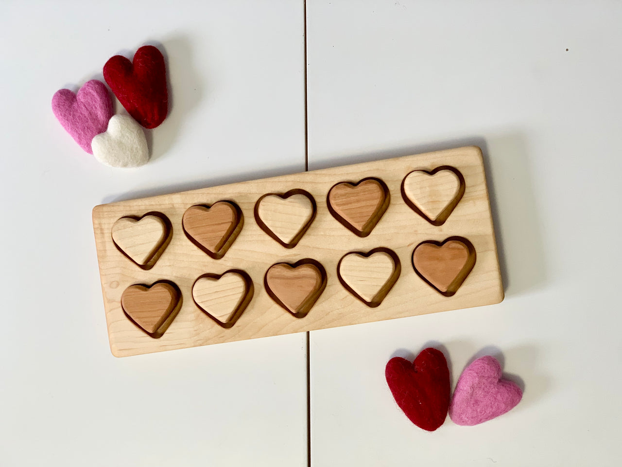 Wooden heart puzzle with alternating light and dark hearts and felt hearts in red, pink, and white nearby on white surface