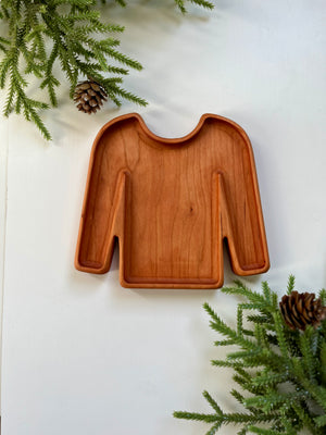 Wooden tray carved in the shape of a long-sleeve sweater surrounded by pine branches and pine cones on white surface