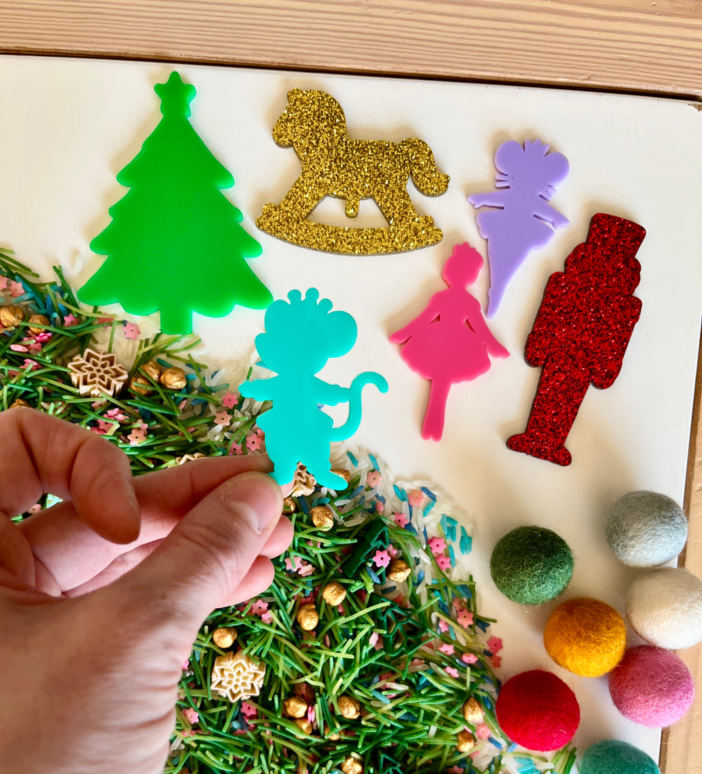 Hand holding blue dinosaur cutout above green, red, purple, pink, and gold holiday-themed acrylic shapes with mixed sensory materials