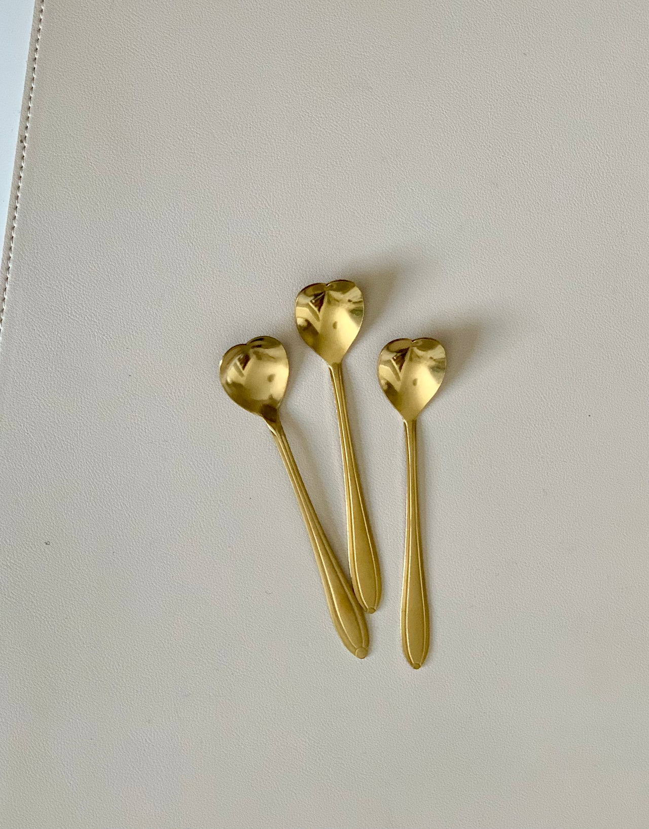 Three gold-toned spoons with heart-shaped bowls arranged on a smooth off-white surface