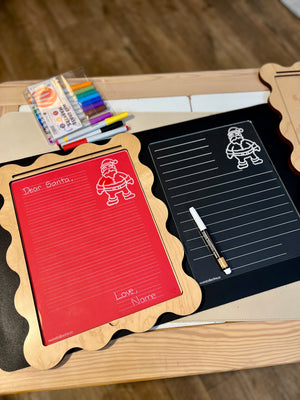 Two wooden-framed writable boards with Santa illustrations, one red with message, one black with white marker, on wooden table with colored markers nearby