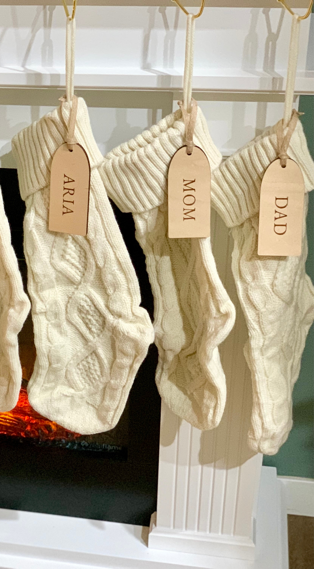 Three cream cable knit Christmas stockings hanging on white mantle with leather name tags labeled Aria, Mom, and Dad