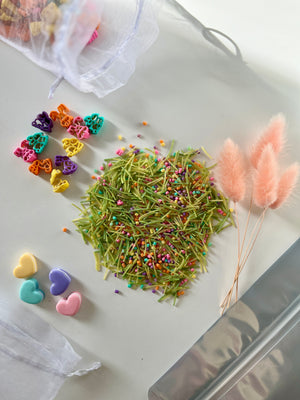 Colorful heart-shaped pasta pieces, green pasta strands with multicolored sprinkles, and pink fluffy dried flowers on white surface