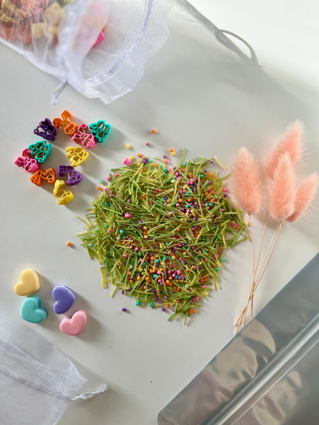 Colorful heart-shaped pasta pieces, green pasta strands with multicolored sprinkles, and pink fluffy dried flowers on white surface