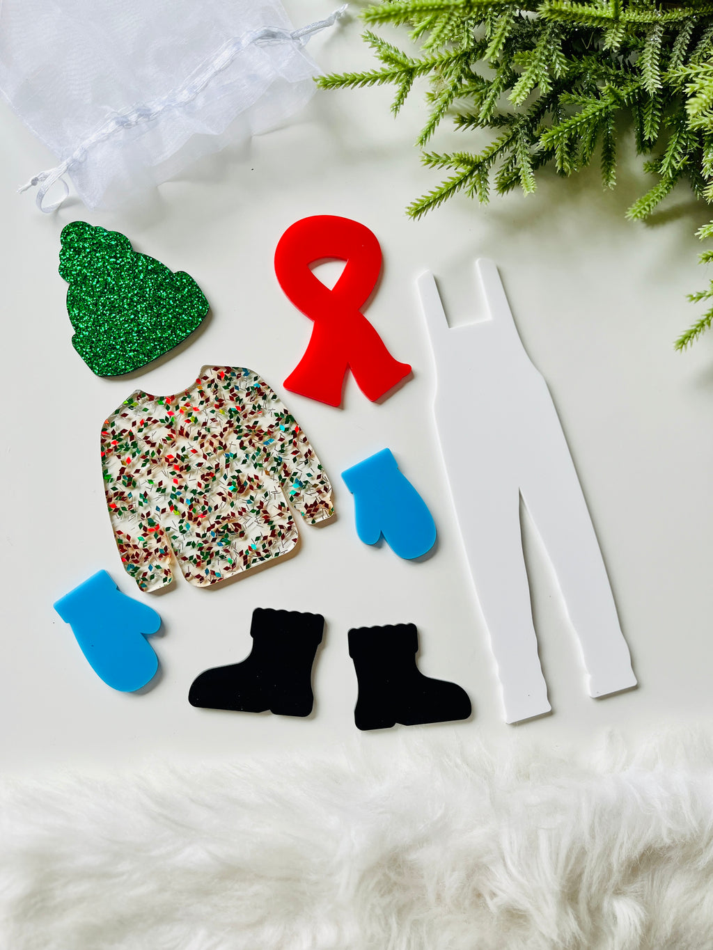 Assortment of acrylic cutouts including white overalls, blue mittens, black boots, red ribbon, glittery green hat, and festive sweater on white surface