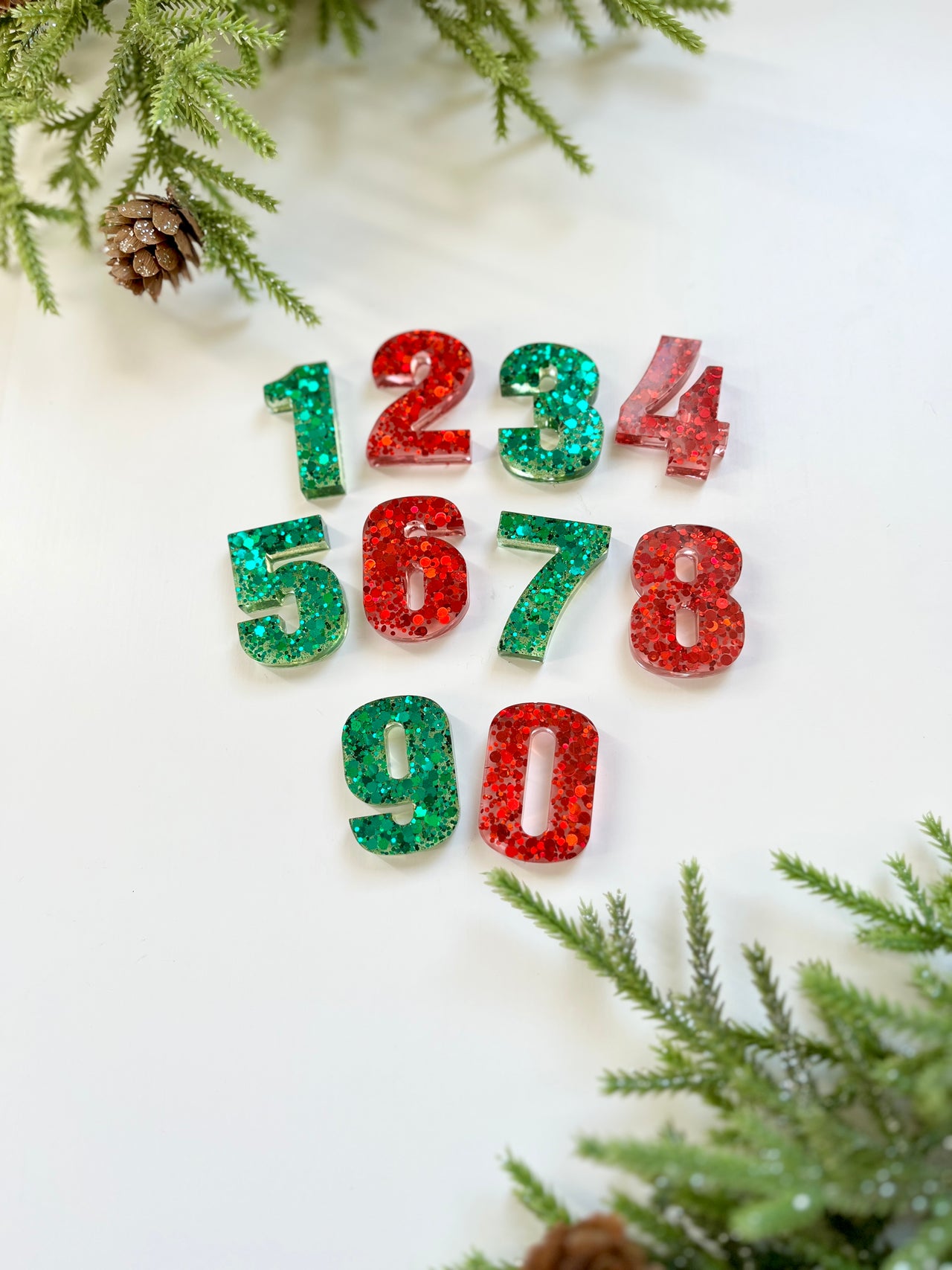Green and red glittery resin numbers arranged on white surface with pine branches and pine cones framing the edges