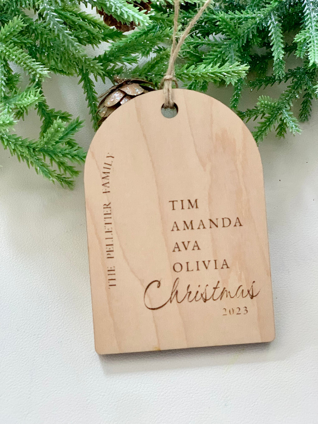 Light wood arch-shaped ornament engraved with family names and "Christmas 2023" hanging from twine against green foliage