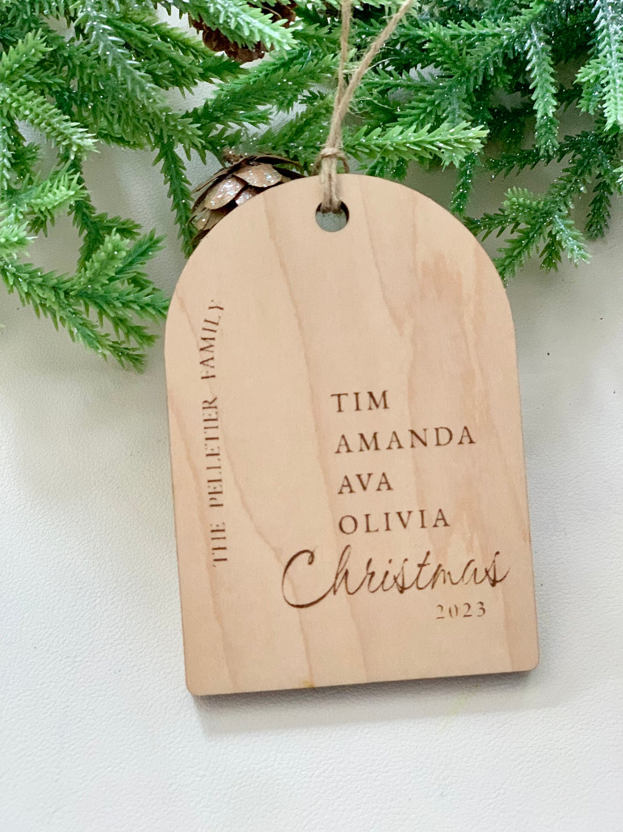 Light wood arch-shaped ornament engraved with family names and "Christmas 2023" hanging from twine against green foliage
