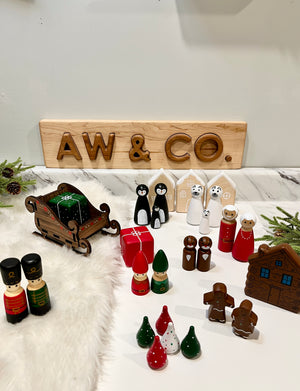 Wooden holiday figurines including toy soldiers, penguins, gingerbread people, wrapped gifts, sleigh, and houses on white surface