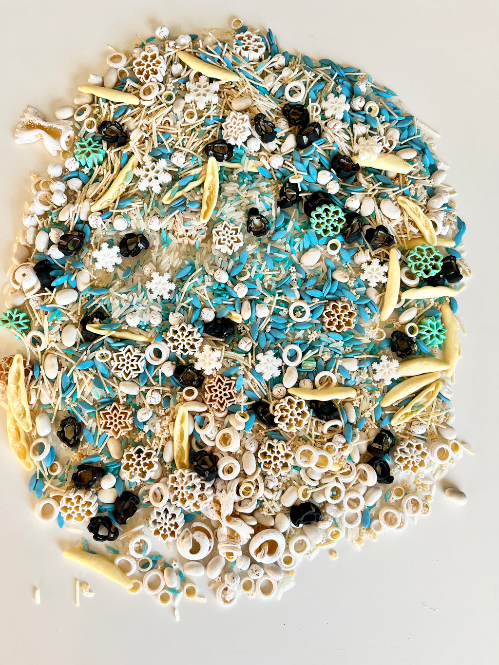 Mixed sensory materials including blue grains, white rings, yellow pods, black seeds, and snowflake shapes on white surface