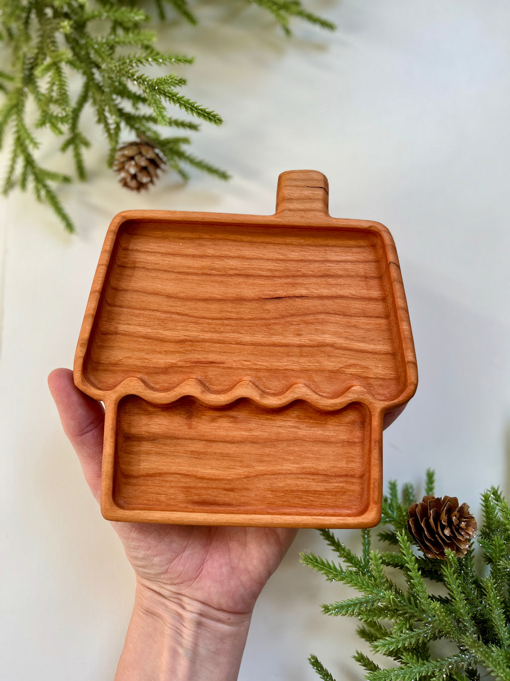 Hand holding a wooden tray shaped like a house with chimney, surrounded by pine branches and pine cones on white surface