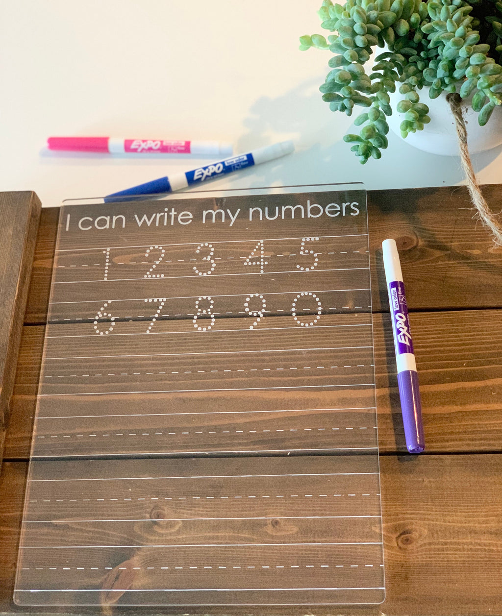 Transparent acrylic number tracing board with dotted digits and three colored dry erase markers on wooden table next to plant