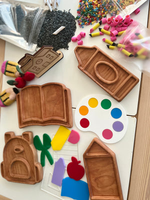 Wooden trays shaped like schoolhouse, book, house, and backpack with colorful craft supplies on light surface