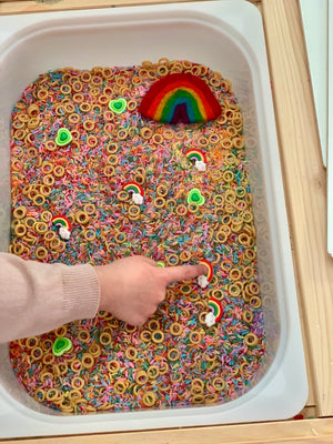 Child’s hand touching rainbow and cloud shapes in sensory bin filled with colorful rice and small ring pieces