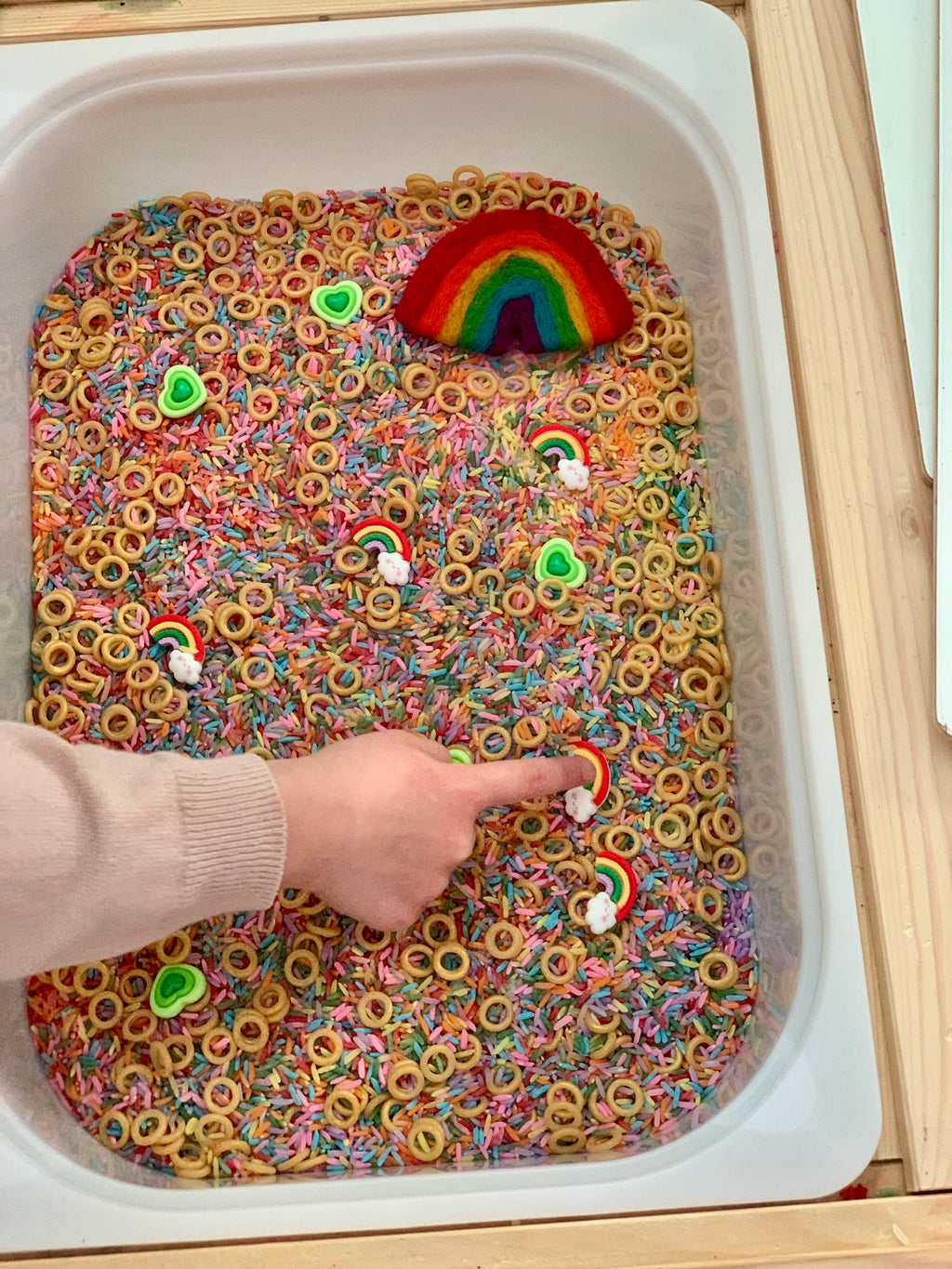 Child’s hand touching rainbow and cloud shapes in sensory bin filled with colorful rice and small ring pieces