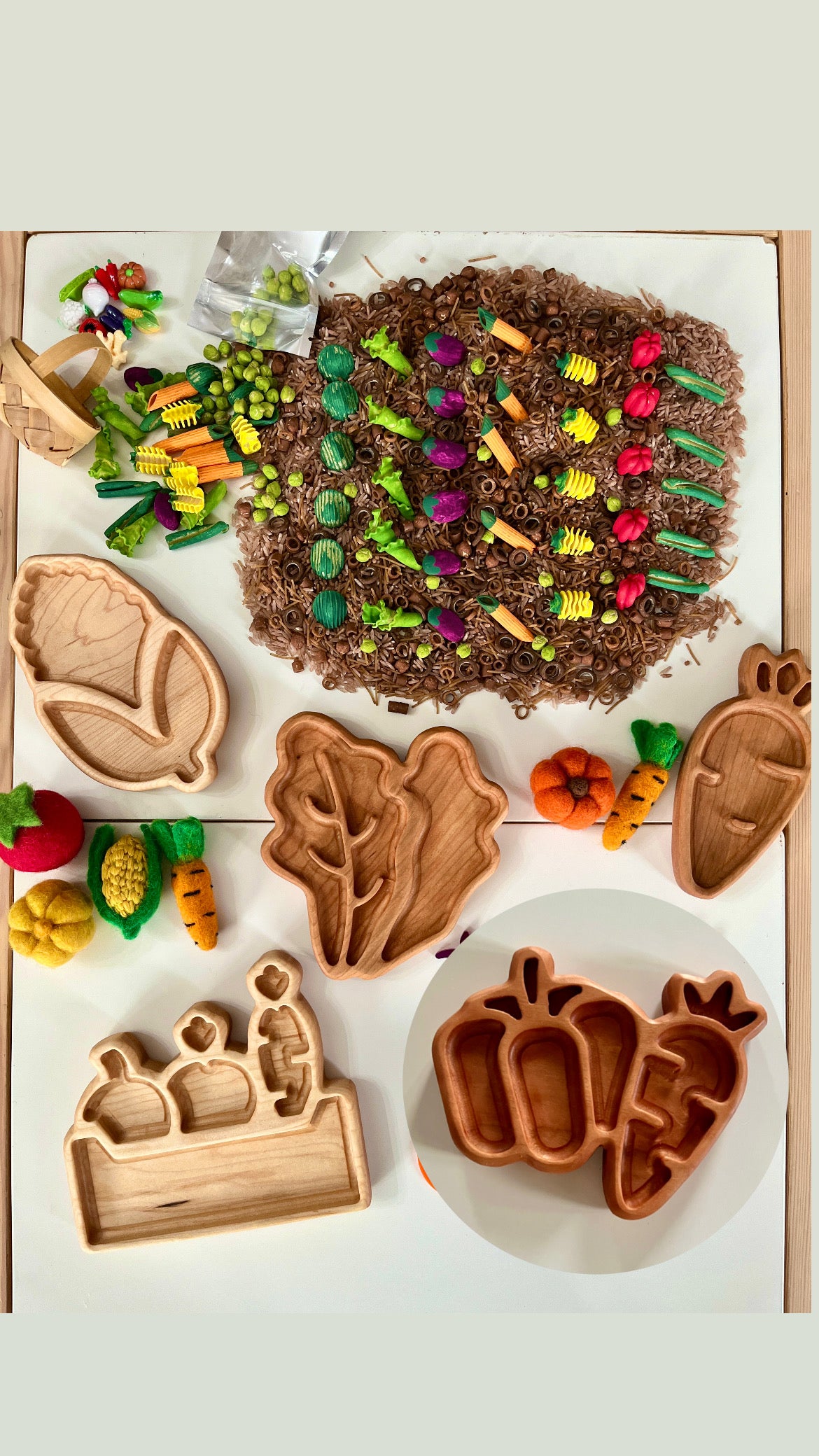 Wooden trays shaped like leaves, vegetables, and garden items surrounded by small felt vegetables and colorful pasta pieces.