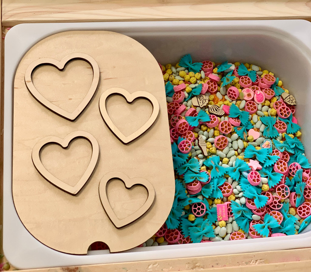 Wooden sensory tray with four heart-shaped cutouts next to bin filled with colorful pasta and dried corn kernels