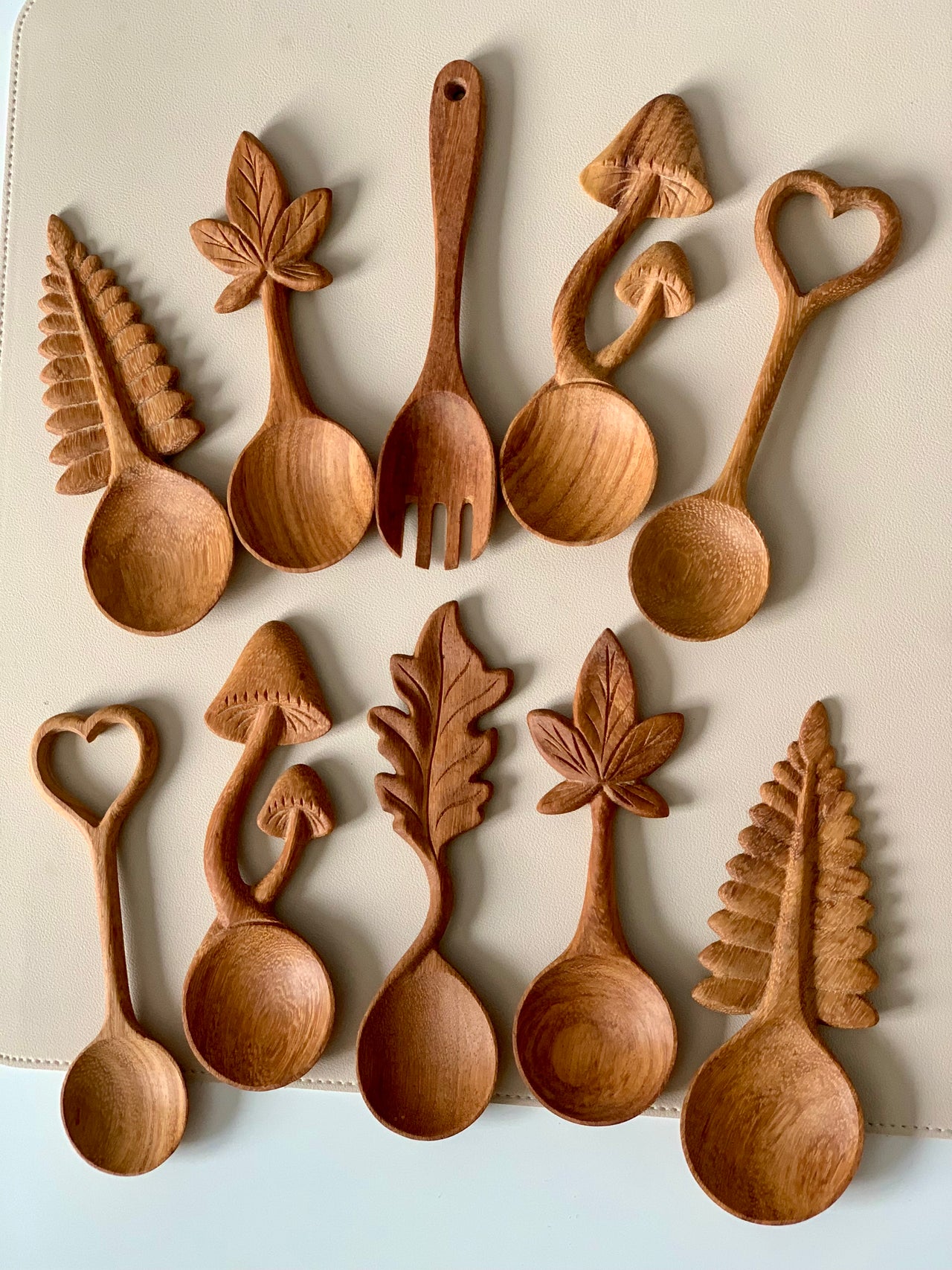 Assortment of carved wooden spoons with handles shaped like leaves, mushrooms, and hearts arranged on a neutral surface