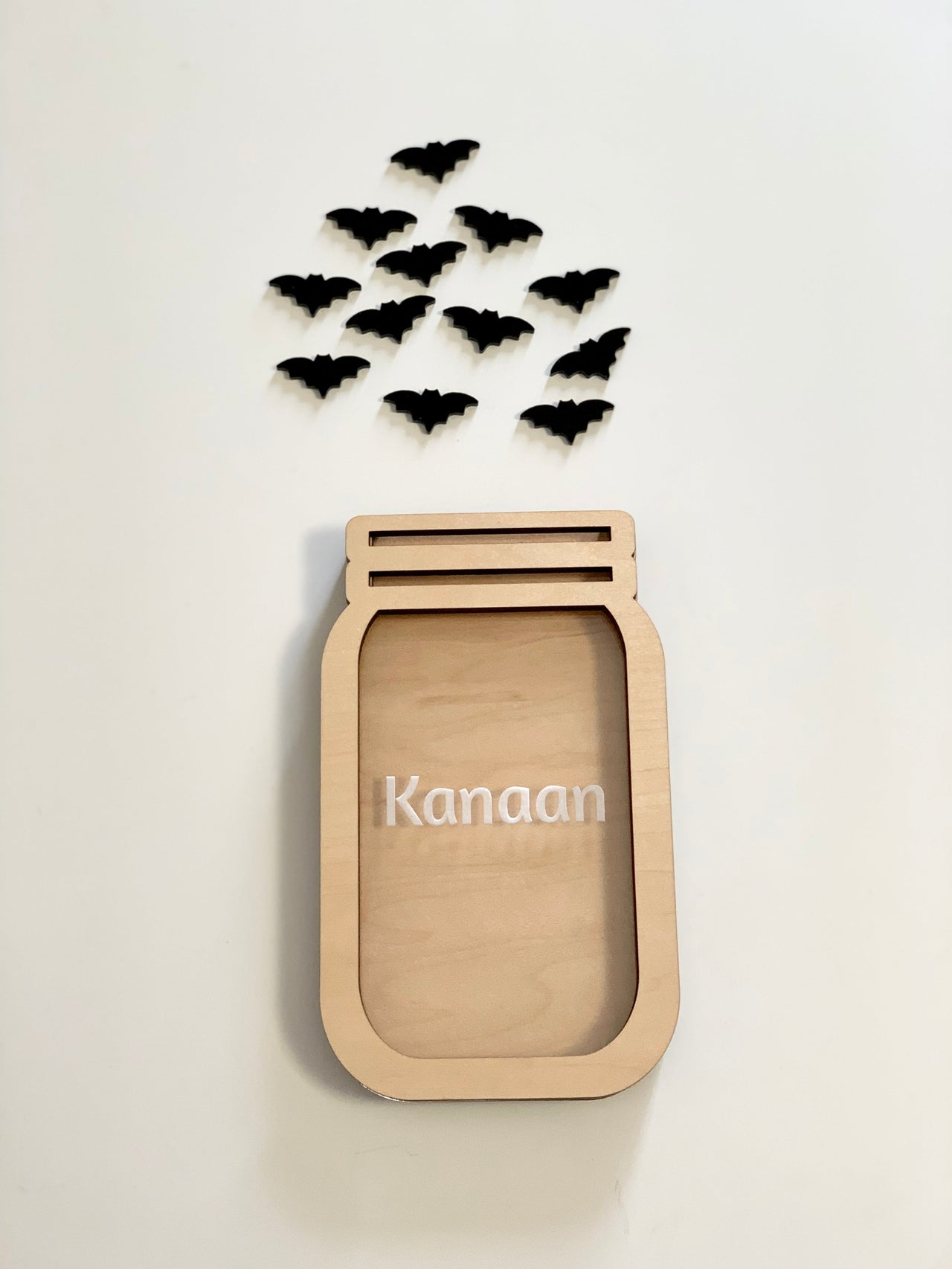 Light wooden jar-shaped frame with the name Kanaan and black bat cutouts arranged above on a white background