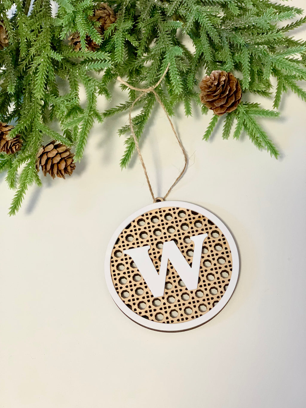Round wooden Christmas ornament with monogram letter W and rattan design hanging near pinecones and green branches