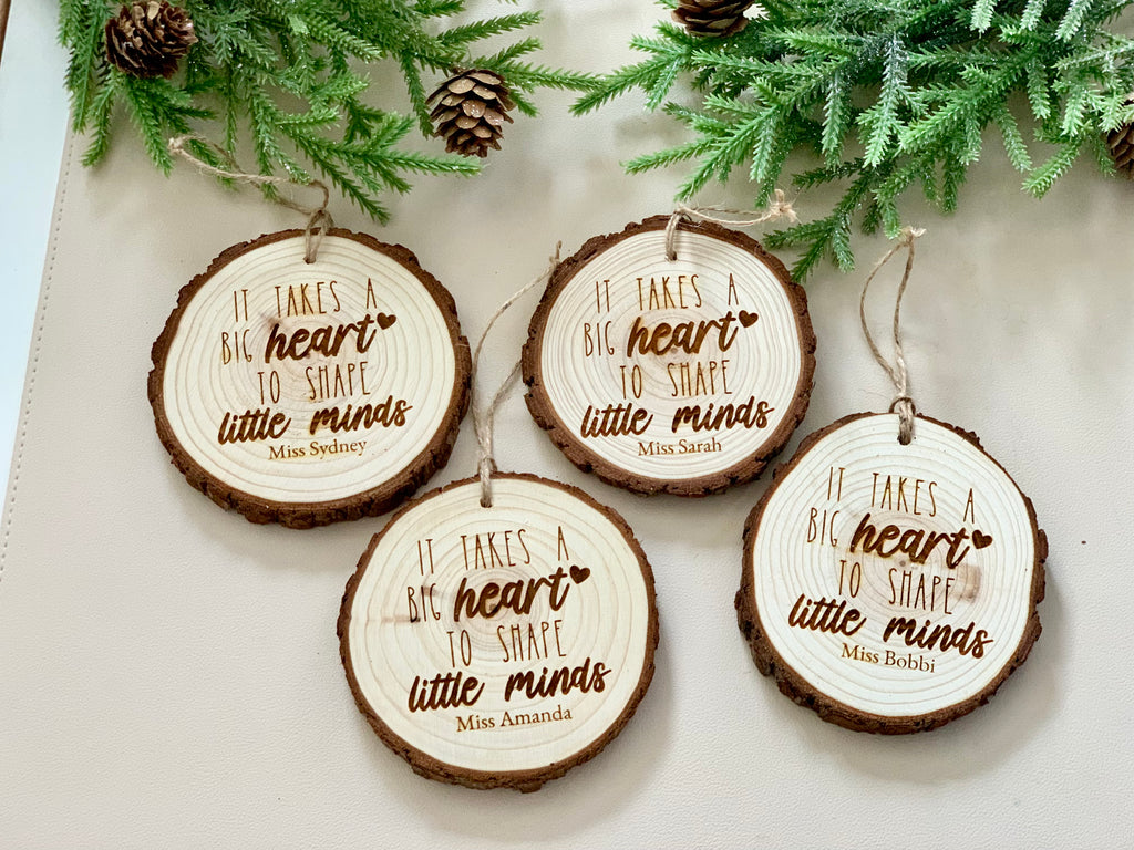 Four round wooden Christmas ornaments with bark edges engraved with "It takes a big heart to shape little minds" and personalized names
