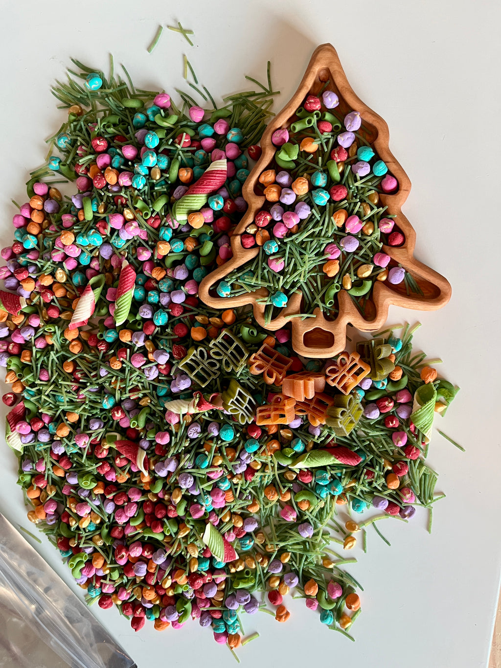 Wooden tree-shaped tray filled with colorful beads, dried green pine needles, and spiral striped pasta on white background