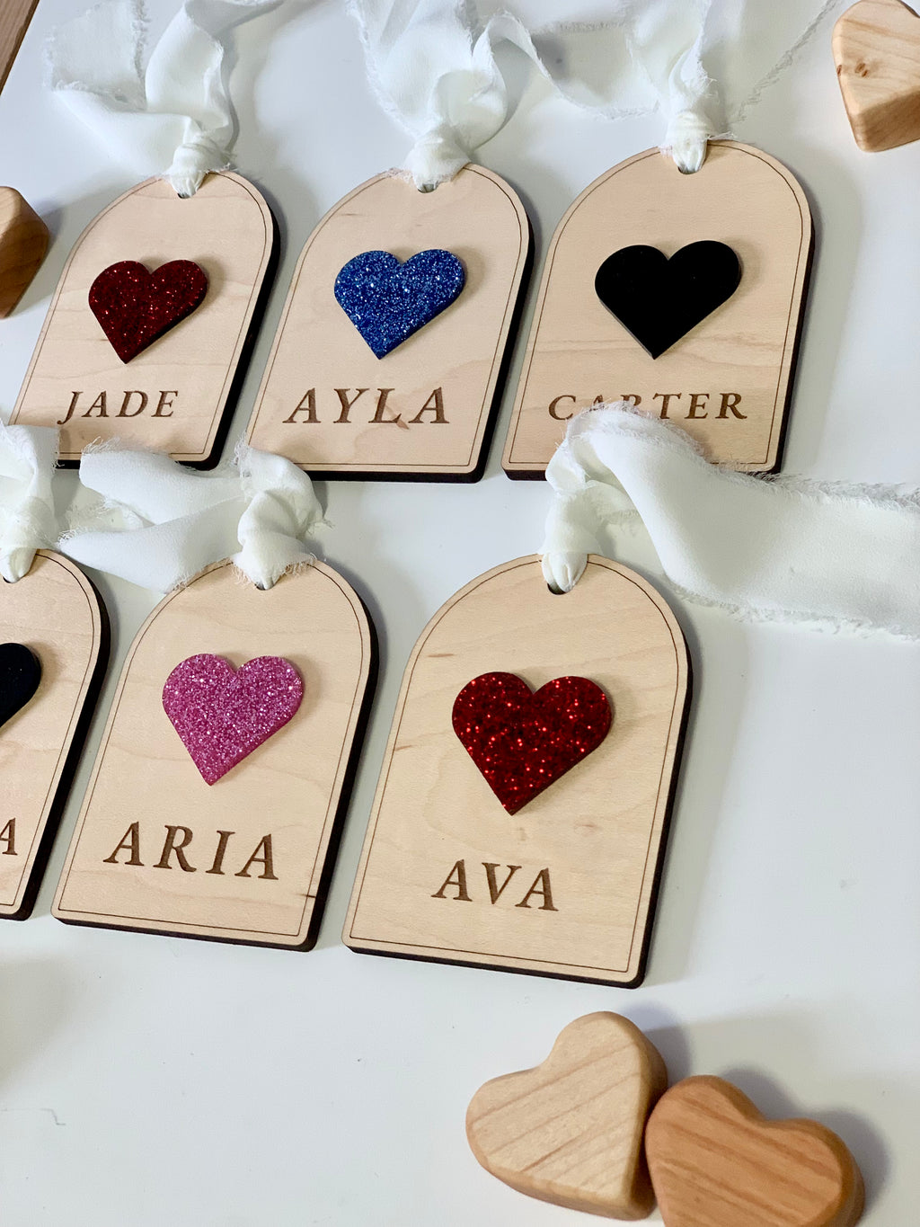Set of wooden gift tags with glittery heart shapes in various colors and engraved names tied with white fabric ribbons