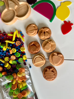 Wooden discs carved with fruit shapes scattered on white surface near colorful pasta pieces and plastic fruit cutouts